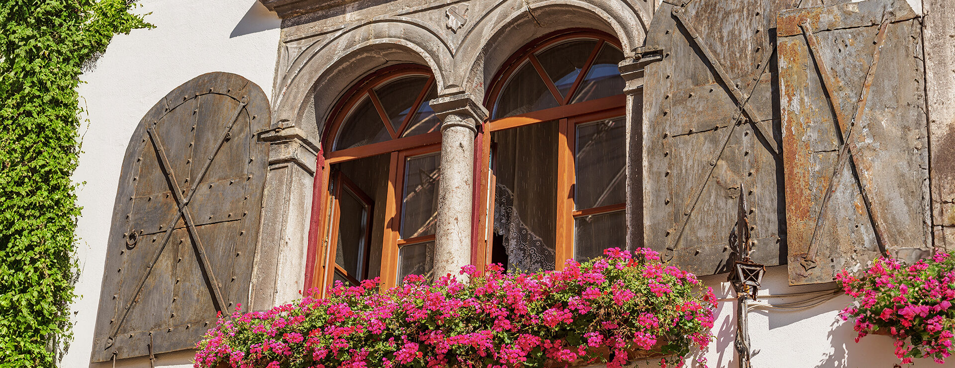 Close-up of an ancient mullion window with metal shutters and geranium flowers. Small village of Malborghetto Valbruna, Val Canale, Udine province, Friuli-Venezia Giulia, Italy, Europe.